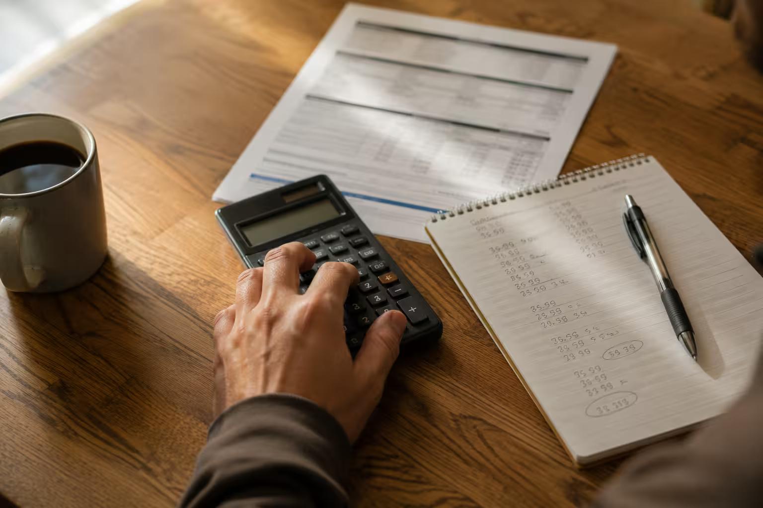 Main sur une calculatrice à côté d'un relevé de prêt automobile et d'un bloc-notes couvert de chiffres, sur une table de bois