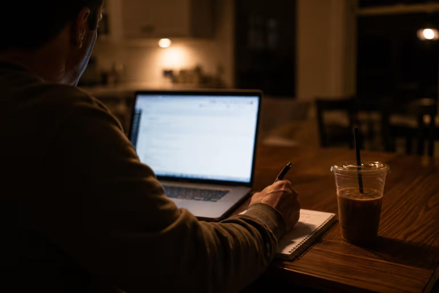 Homme comparant des offres de financement automobile sur son ordinateur portable, tard le soir à la table de cuisine, en prenant des notes